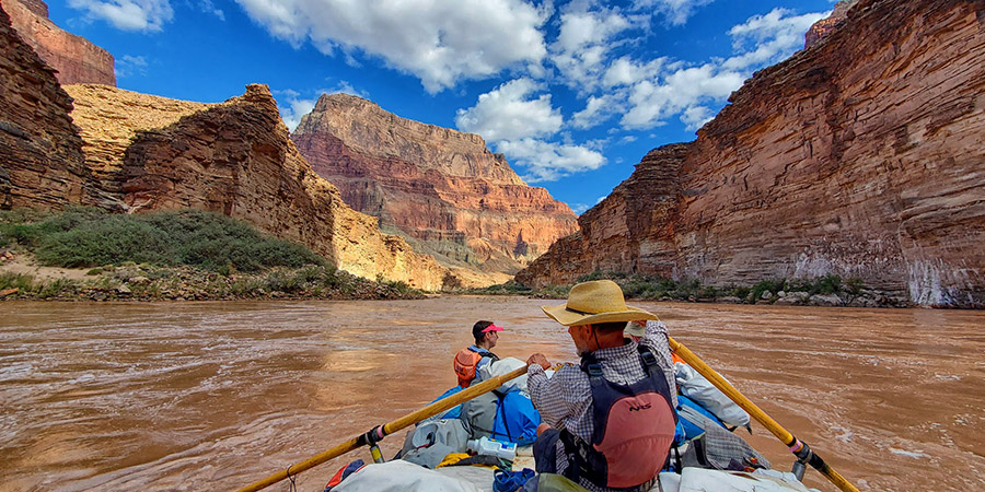 Boatman leans back, rowing a muddy Colorado River in the Grand Canyon under a brilliant blue sky with wispy white clouds