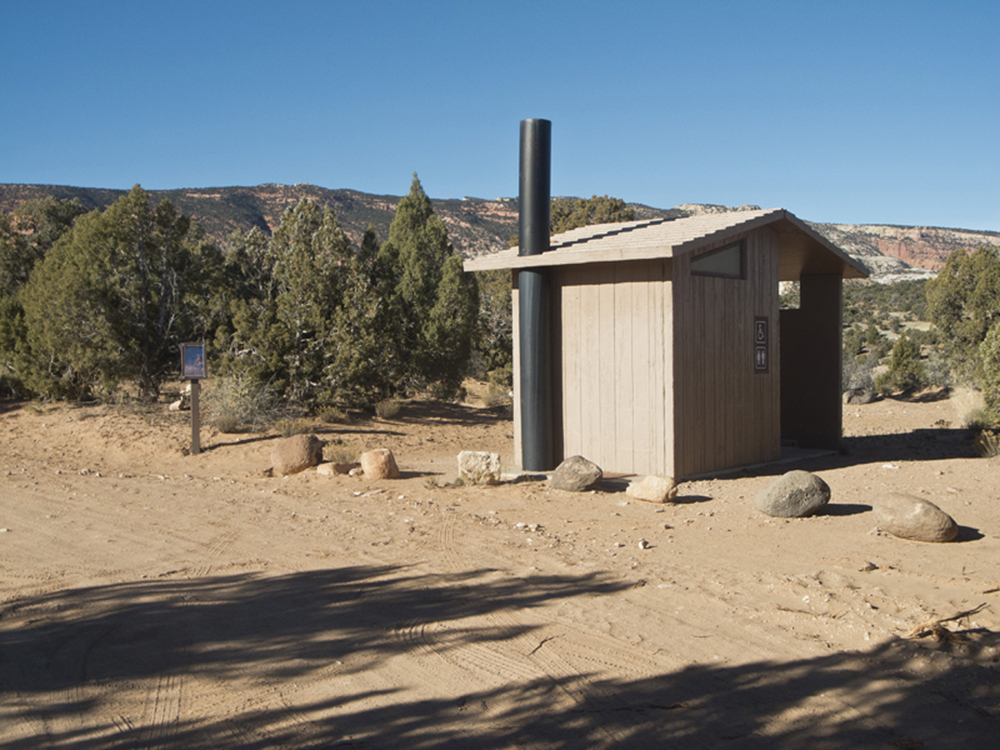 A grey building with a vault toilet at Cedar Mesa Campground