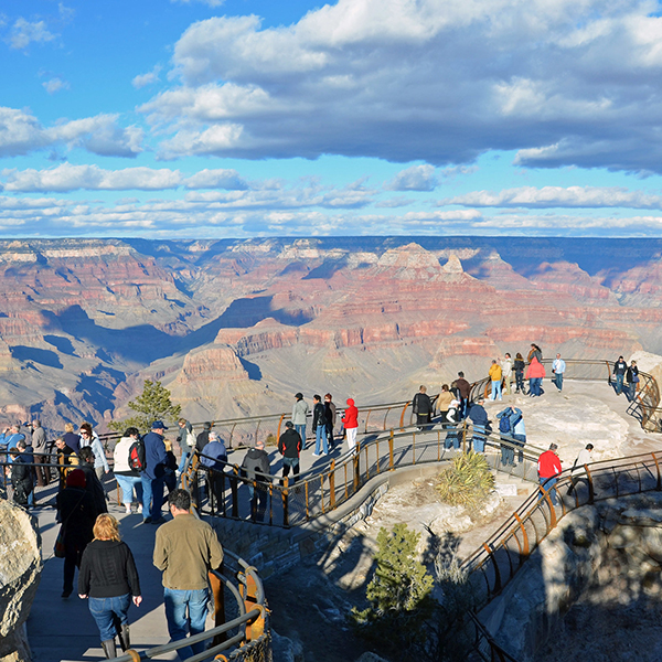 A crow of people in brightly colored clothes stands at the handrail looking at the Grand Canyon