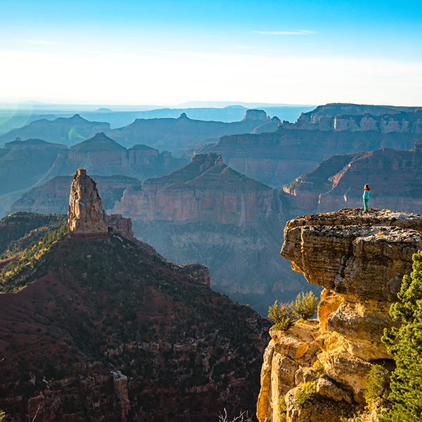 A lone woman in bright clothing stands on a rock outcropping looking out at the grand canyon