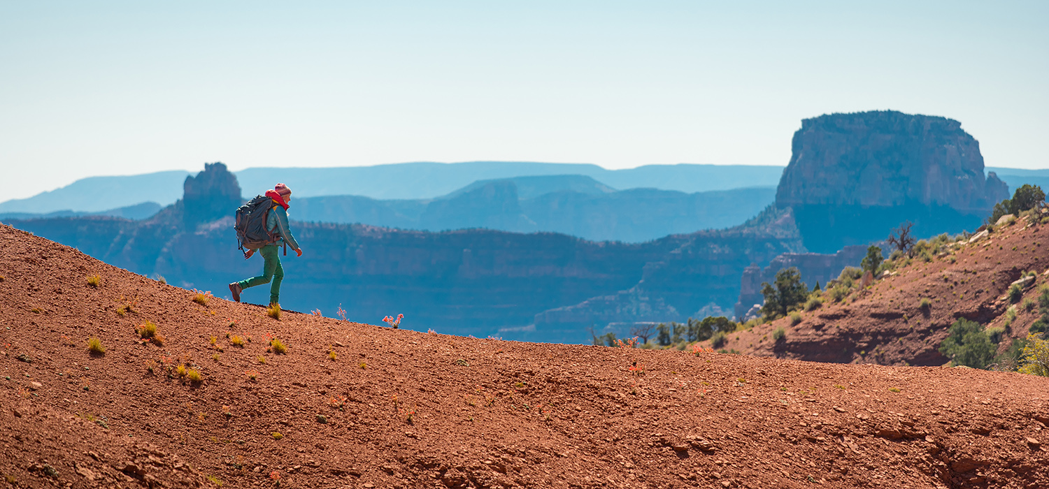 A hiker in bring clothes walks along orange rocks on the north rim of the grand canyon, which looks blue in the distance