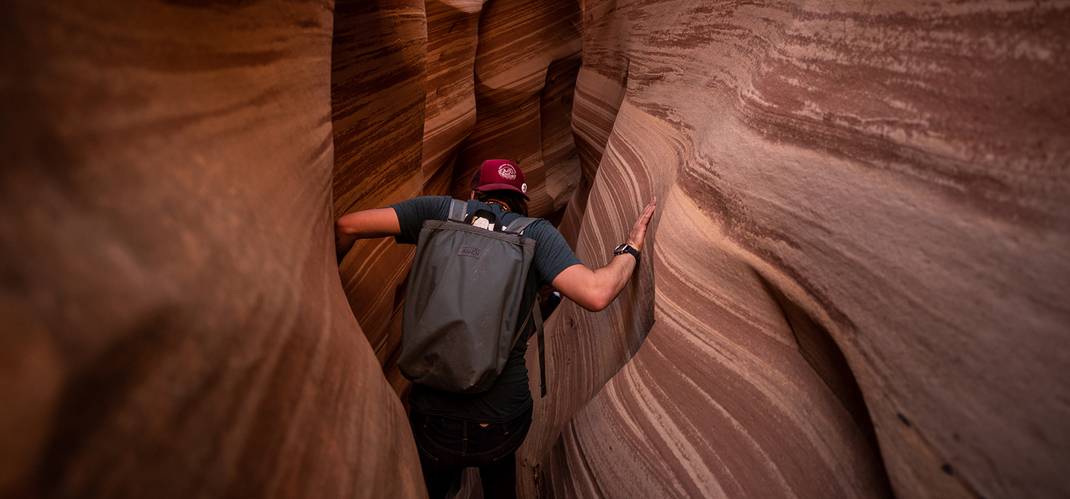 A hiker in a red hat crawls through a slot canyon whose walls are reddish with white stripes