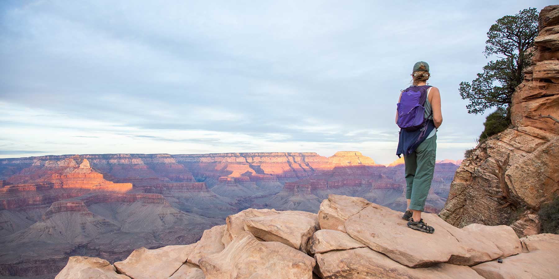 Woman in a purple backpack stands looking out at the Grand Canyon