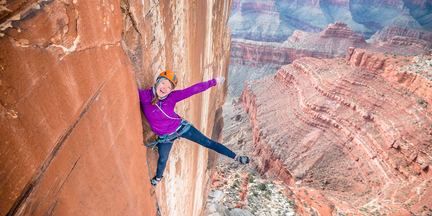 Smiling woman in bright pink shirt with her arm and leg outstretched, rock climbing in the Grand Canyon