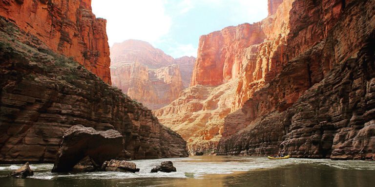 Yellow boat on glassy water in the Colorado River surrounded by Grand Canyon cliffs which go from dark brown to bright orange, like a painting