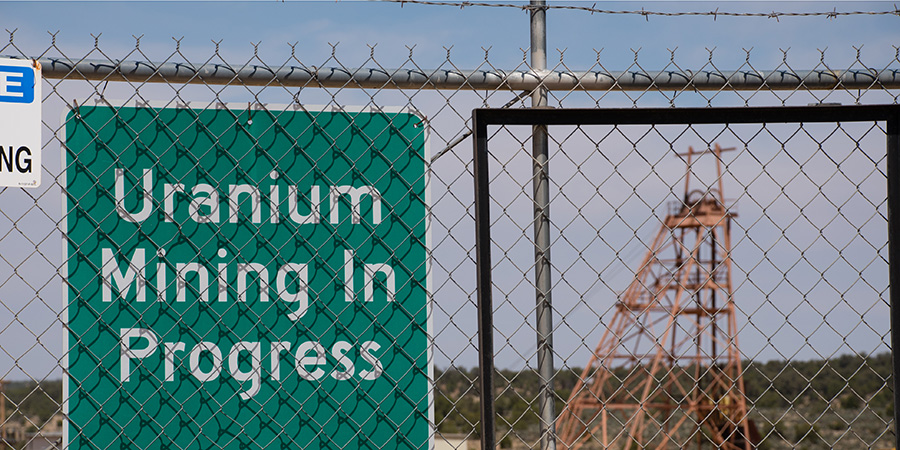 Green sign with white lettering reading Uranium Mining in Progress on the chainlink fence at Canyon Mine, renamed Pinyon Plain Mine, a uranium mine near the Grand Canyon