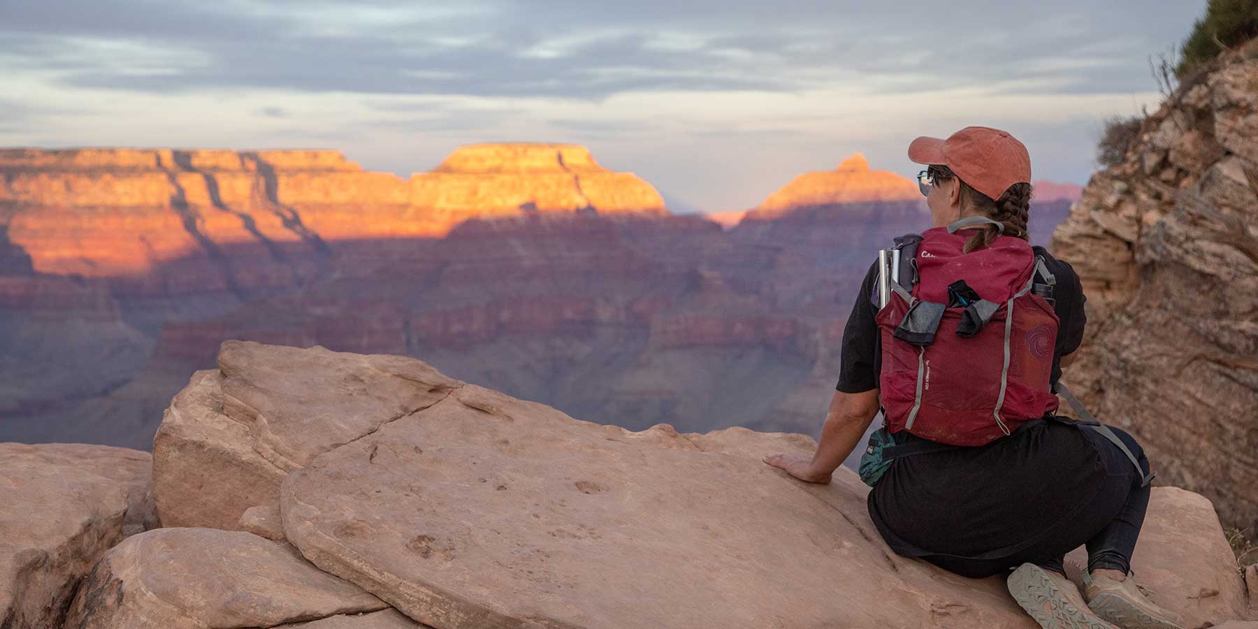 A hiker wearing a red backpack and orange ball cap perches on a sandstone rock, looking out at the Grand Canyon