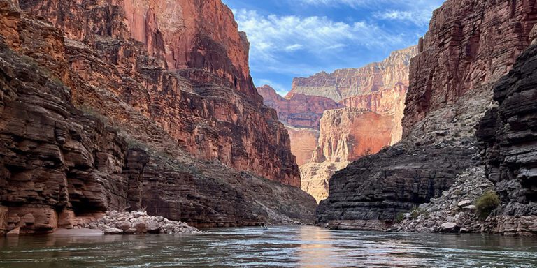 Clear calm green Colorado River with step red, pink, brown, and orange Grand Canyon cliffs on both sides.