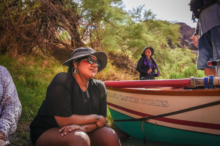 Young Native woman in sunglasses and hat sits beside a dory in on the bank of the Colorado River in the grand canyon