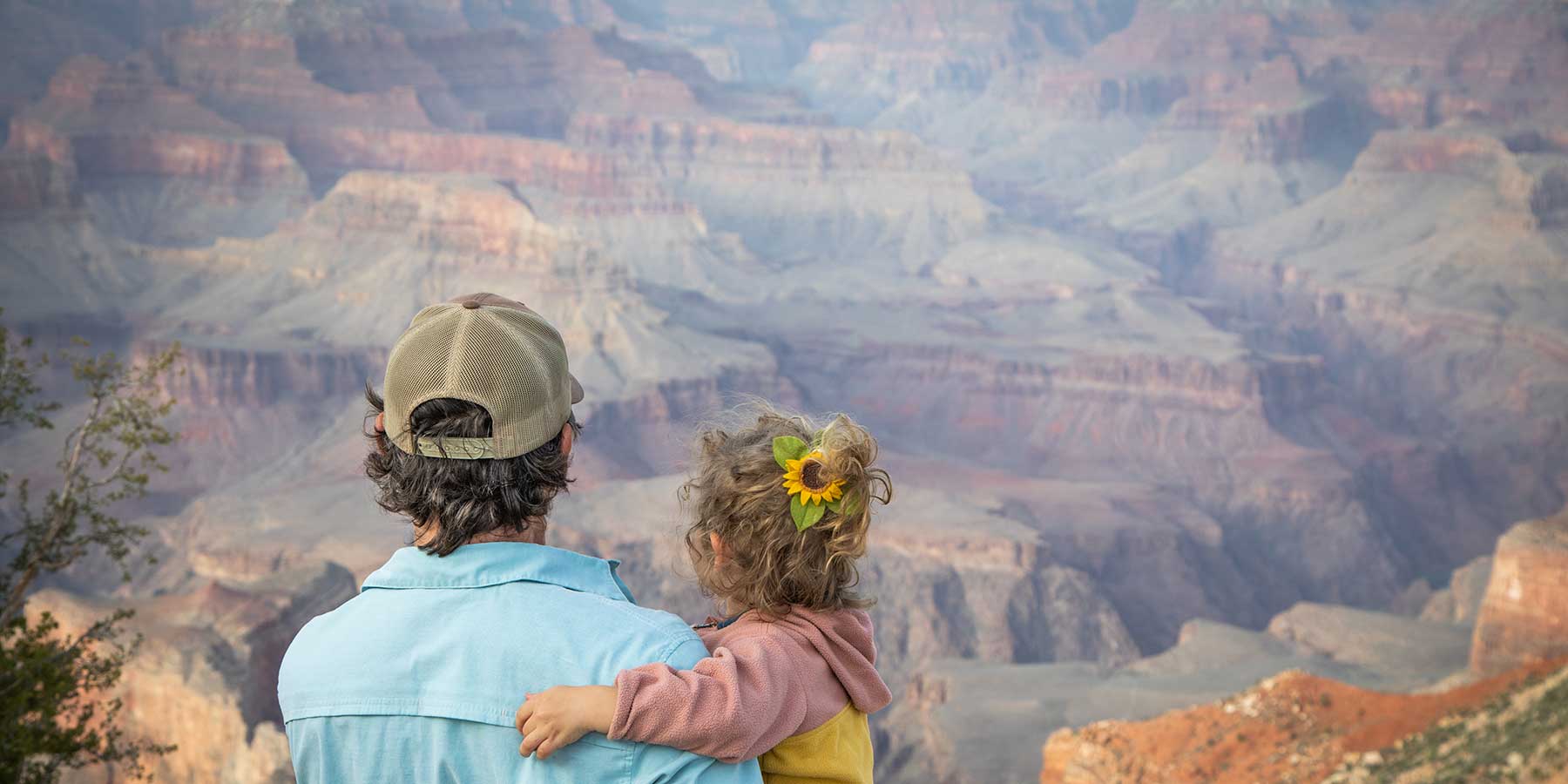 A man holds a young girl with a sunflower in her hair and looks out at the Grand Canyon