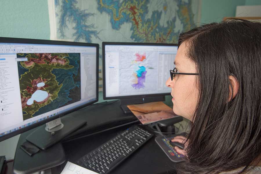 A woman wearing glasses works on her computer with maps displaying.