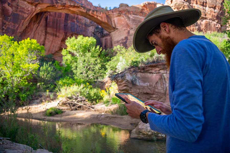 A man wearing a hat and red beard enters data on an ipad in the Escalante River cooridor.