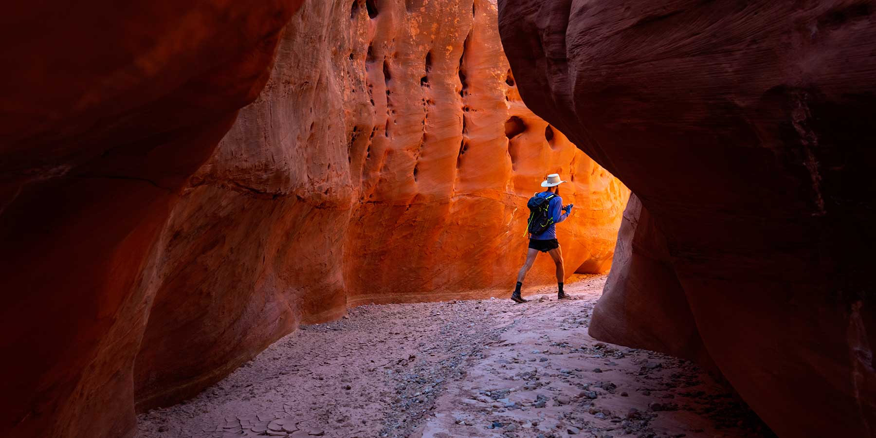 A man in a hat rounds a bend in a red slot canyon walking toward golden light