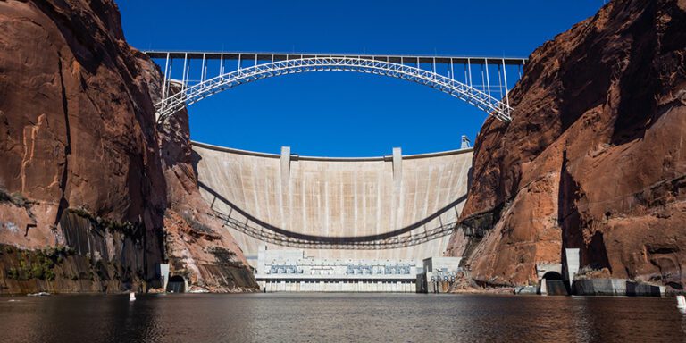 Looking up at Glen Canyon Dam from the Colorado River, including cliffs, large concrete dam and a bridge overhead