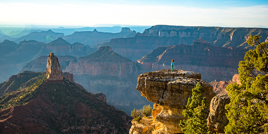 Woman in bright clothes stands on a rock outcropping looking out at the Grand Canyon whose rock layers fade to blue in the distance