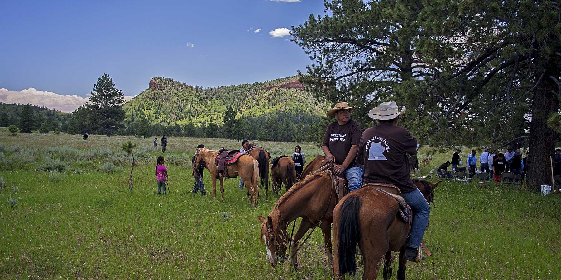 Two men on horseback in Protect Bears Ears t-shirts with more riders in the distance in a meadow below the Bears Ears buttes