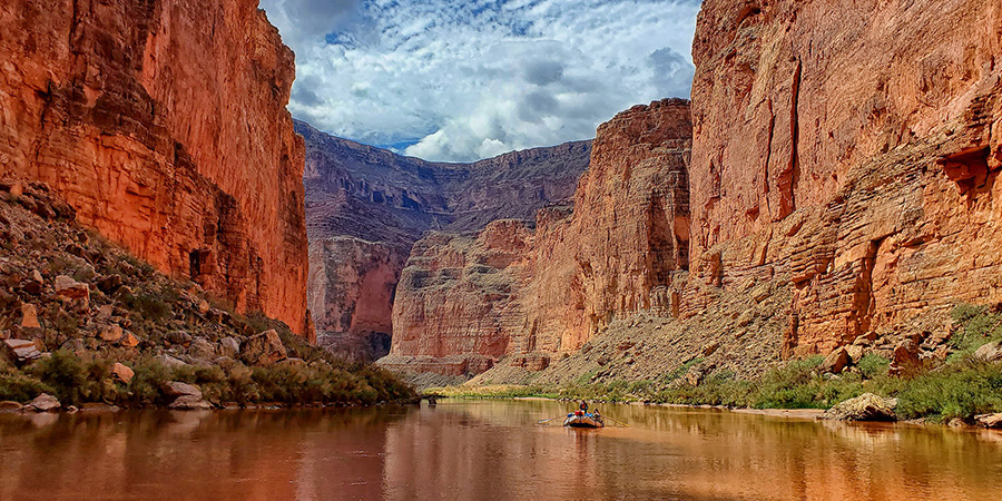 An inflatable boat rows down the shiny flat muddy Colorado River within Marble Canyon orange cliffs