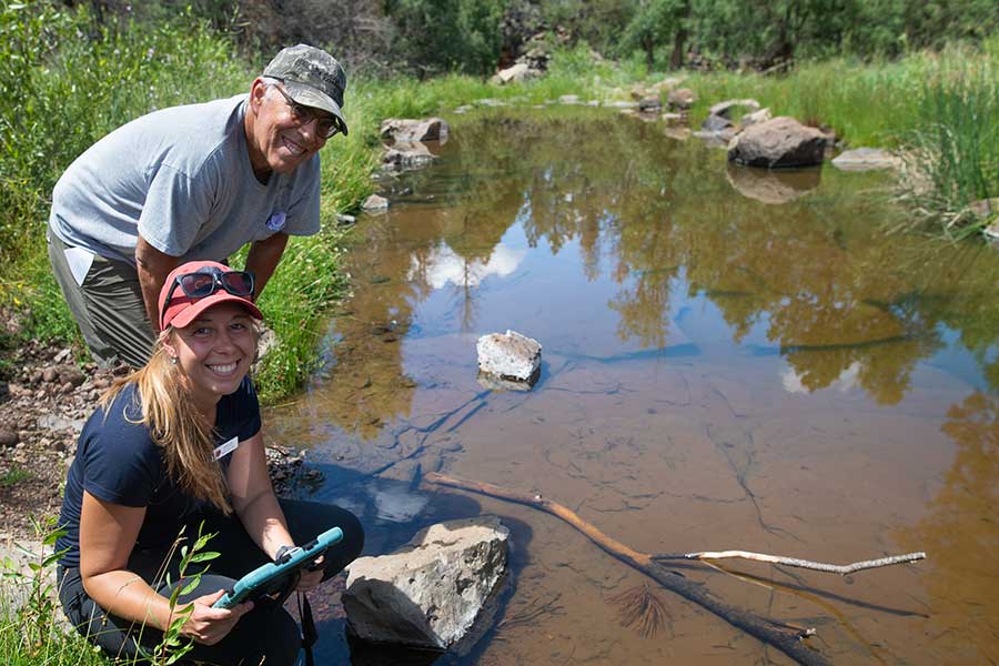 Two volunteers smile while recording data at a spring in Arizona, beside a pool of clear water surrounded by plants