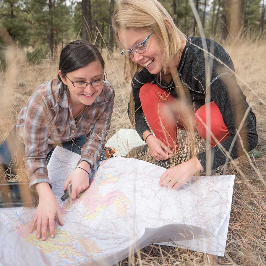 Two women, one with blonde hair one with black hair, look at a field map in the forest.