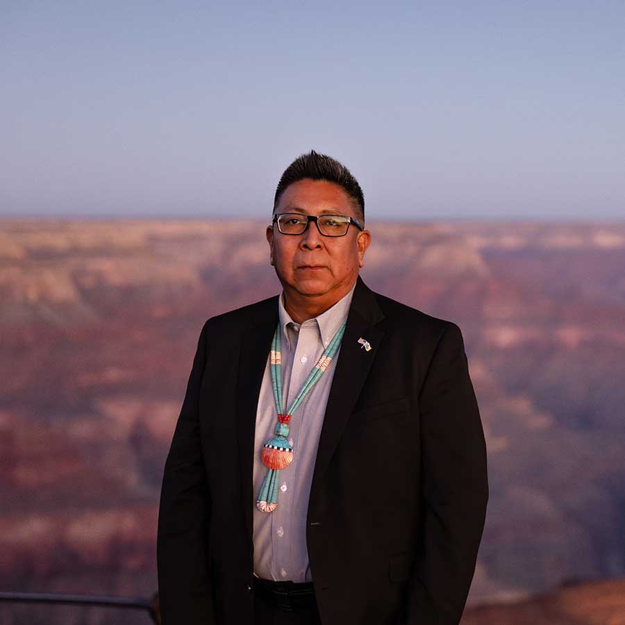 Timothy Nuvangyaoma, of the Hopi Tribe, in a black suit jacket, white shirt, and necklace with the pink rock layers of the Grand Canyon in the background