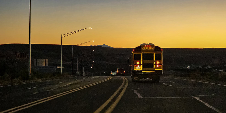 A school bus with its brake lights on rounding a bend at dusk behind trucks on Highway 160, along the uranium haul route