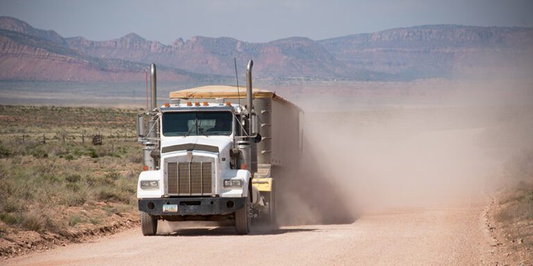 White uranium haul truck covered with a tarp drives along a dirt road in a cloud of dust.