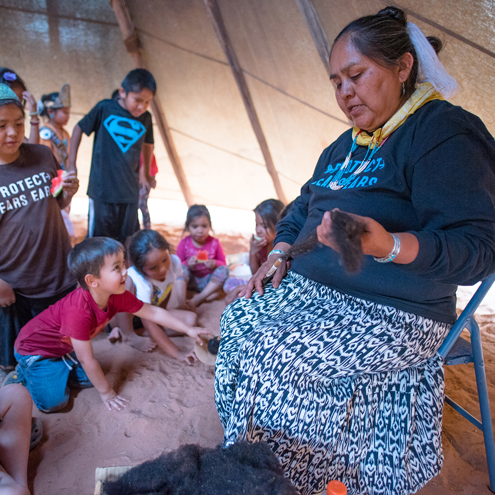 A Native woman sits in a chair and speaks to children assembled at her feet inside a teepee