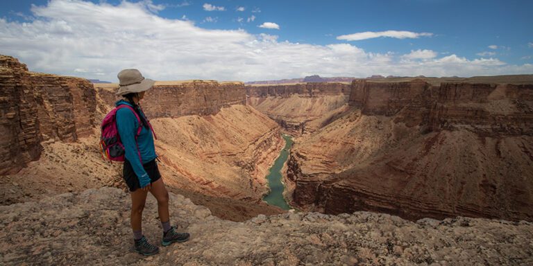 A person in hiking attire stands on a rocky cliff within Baaj Nwaavjo I'tah Kukveni Grand Canyon national monument, overlooking a vast canyon where a river winds through under a partly cloudy sky.