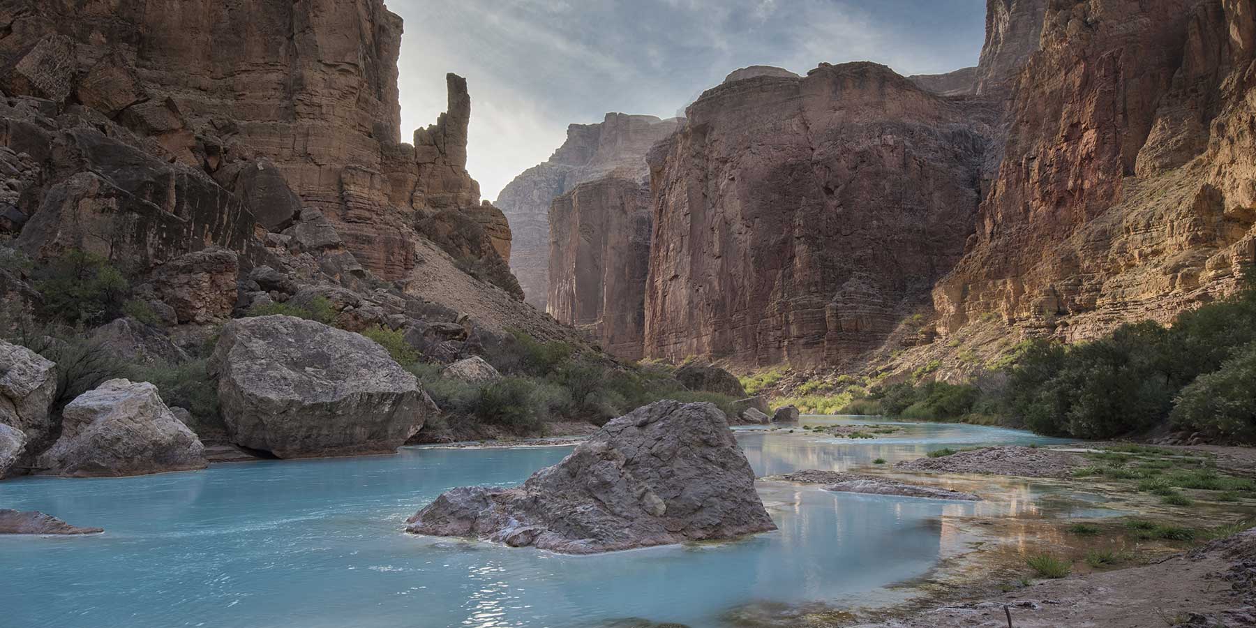 Milky turquoise river flows around large rock with high reddish cliffs on both sides, the Little Colorado River near the Grand Canyon.