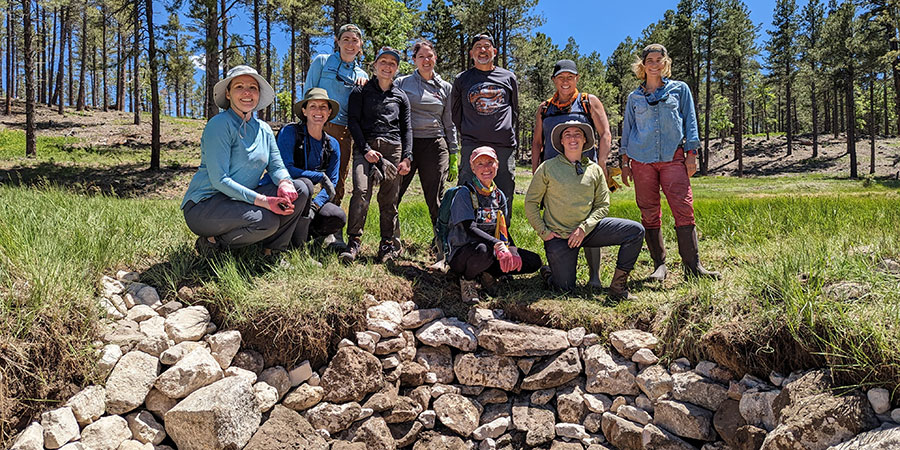 A group of people outdoors, standing and kneeling near a rock-lined area, with trees and grass in the background.