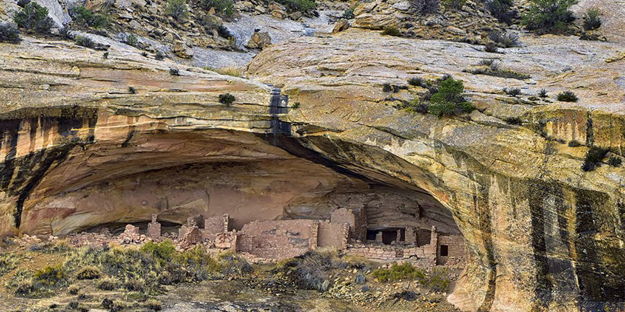 An ancient cliff dwelling with stone structures set beneath a large, curved rock formation, surrounded by the sparse vegetation and rocky terrain, Butler Alcove, Bears Ears National Monument