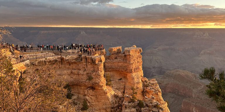 As the sun dips below the horizon, casting a warm glow over the Grand Canyon, visitors savor every moment of their one-day adventure. They stand atop a rocky overlook, mesmerized by the clouds and stunning layered canyon scenery stretching far into the distance.